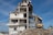 A partially demolished building with exposed beams, debris surrounding the base, and an excavator actively working on the site. The structure shows severe damage with torn walls and collapsed sections. The sky is clear and blue.