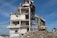 A partially demolished building with exposed beams, debris surrounding the base, and an excavator actively working on the site. The structure shows severe damage with torn walls and collapsed sections. The sky is clear and blue.