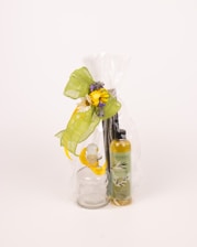 A beautifully wrapped gift set featuring a clear glass bottle with a stopper, a bottle of scented oil labeled 'Esteban Paris', and a decorative arrangement that includes green ribbon, small flowers, and a yellow bow. The items are contained in a transparent plastic wrap.