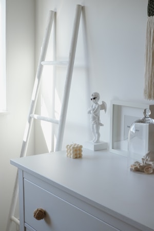 A minimalistic and modern interior setting includes a white dresser with a decorative knob and various items on top. A white sculptural angel figurine wearing sunglasses stands on the dresser, next to a candle shaped like a cluster of spheres. A glass dome covers a small arrangement of shells or coral. A tall white ladder leans against the wall, and a macrame wall hanging is partially visible.