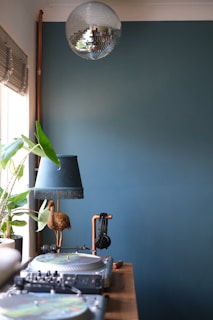 A room featuring a DJ setup with turntables and a mixer on a wooden table. A pair of headphones is hanging from a copper stand. There is a decorative lamp with a green shade and a golden bird sculpture. To the left, a potted houseplant sits on a windowsill with blinds partially drawn. A silver disco ball hangs from the ceiling. The background wall is painted a muted teal color.