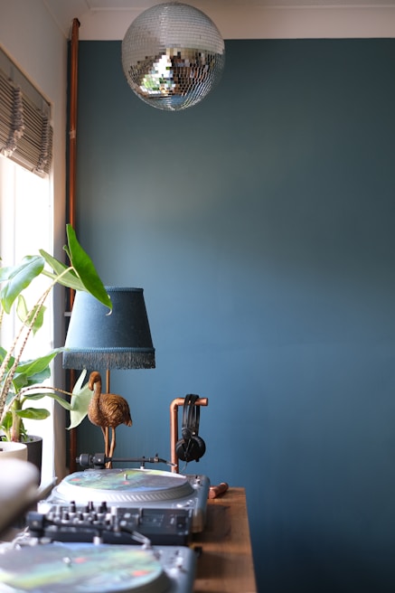 A room featuring a DJ setup with turntables and a mixer on a wooden table. A pair of headphones is hanging from a copper stand. There is a decorative lamp with a green shade and a golden bird sculpture. To the left, a potted houseplant sits on a windowsill with blinds partially drawn. A silver disco ball hangs from the ceiling. The background wall is painted a muted teal color.