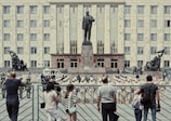 A large building serves as the backdrop for a military parade, with soldiers in uniform standing in formation in front of a tall statue. Civilians stand behind a fence observing the event. The statues on either side of the central monument depict historical figures in dynamic poses.