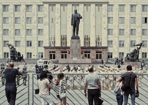 A large building serves as the backdrop for a military parade, with soldiers in uniform standing in formation in front of a tall statue. Civilians stand behind a fence observing the event. The statues on either side of the central monument depict historical figures in dynamic poses.