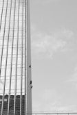 Three individuals are suspended on ropes, cleaning or maintaining the glass facade of a tall building. The building features a vertical, grid-like design with numerous windows. The sky is clear, contributing to the stark contrast and emphasizing the height of the structure.