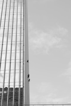 Three individuals are suspended on ropes, cleaning or maintaining the glass facade of a tall building. The building features a vertical, grid-like design with numerous windows. The sky is clear, contributing to the stark contrast and emphasizing the height of the structure.