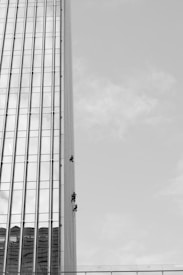 Three individuals are suspended on ropes, cleaning or maintaining the glass facade of a tall building. The building features a vertical, grid-like design with numerous windows. The sky is clear, contributing to the stark contrast and emphasizing the height of the structure.