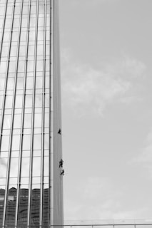 Three individuals are suspended on ropes, cleaning or maintaining the glass facade of a tall building. The building features a vertical, grid-like design with numerous windows. The sky is clear, contributing to the stark contrast and emphasizing the height of the structure.