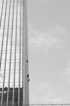 Three individuals are suspended on ropes, cleaning or maintaining the glass facade of a tall building. The building features a vertical, grid-like design with numerous windows. The sky is clear, contributing to the stark contrast and emphasizing the height of the structure.
