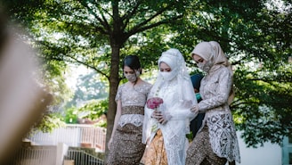A bride dressed in white lace and holding a bouquet of flowers is accompanied by two women in traditional attire. They are walking outdoors surrounded by lush green trees, creating a serene and intimate atmosphere.