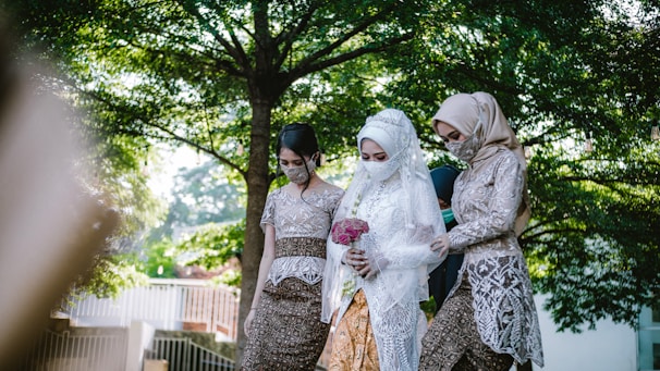 A bride dressed in white lace and holding a bouquet of flowers is accompanied by two women in traditional attire. They are walking outdoors surrounded by lush green trees, creating a serene and intimate atmosphere.