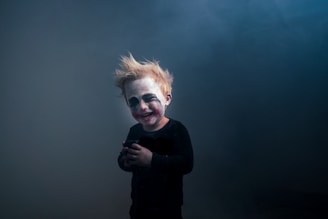 A child with theatrical makeup resembling a clown, featuring exaggerated eyes and mouth marks. The child has messy blonde hair and is wearing a dark outfit, standing against a smoky, dimly lit background.