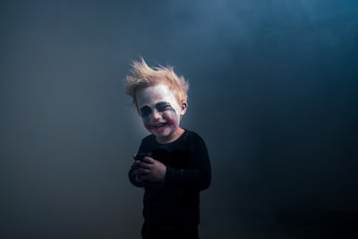 A child with theatrical makeup resembling a clown, featuring exaggerated eyes and mouth marks. The child has messy blonde hair and is wearing a dark outfit, standing against a smoky, dimly lit background.