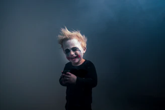 A child with theatrical makeup resembling a clown, featuring exaggerated eyes and mouth marks. The child has messy blonde hair and is wearing a dark outfit, standing against a smoky, dimly lit background.