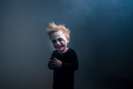 A child with theatrical makeup resembling a clown, featuring exaggerated eyes and mouth marks. The child has messy blonde hair and is wearing a dark outfit, standing against a smoky, dimly lit background.