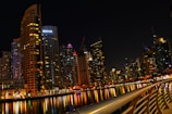 A vibrant cityscape at night with towering skyscrapers that are brightly lit, reflecting on a calm body of water. The skyline includes a variety of modern architectural designs, and a prominent building with the logo 'EMAAR' is visible. A curved railing in the foreground adds depth and perspective to the scene.