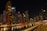 A vibrant cityscape at night with towering skyscrapers that are brightly lit, reflecting on a calm body of water. The skyline includes a variety of modern architectural designs, and a prominent building with the logo 'EMAAR' is visible. A curved railing in the foreground adds depth and perspective to the scene.