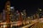 A vibrant cityscape at night with towering skyscrapers that are brightly lit, reflecting on a calm body of water. The skyline includes a variety of modern architectural designs, and a prominent building with the logo 'EMAAR' is visible. A curved railing in the foreground adds depth and perspective to the scene.