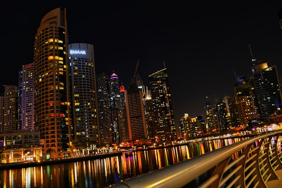 A vibrant cityscape at night with towering skyscrapers that are brightly lit, reflecting on a calm body of water. The skyline includes a variety of modern architectural designs, and a prominent building with the logo 'EMAAR' is visible. A curved railing in the foreground adds depth and perspective to the scene.