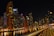 A vibrant cityscape at night with towering skyscrapers that are brightly lit, reflecting on a calm body of water. The skyline includes a variety of modern architectural designs, and a prominent building with the logo 'EMAAR' is visible. A curved railing in the foreground adds depth and perspective to the scene.