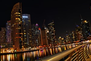 A vibrant cityscape at night with towering skyscrapers that are brightly lit, reflecting on a calm body of water. The skyline includes a variety of modern architectural designs, and a prominent building with the logo 'EMAAR' is visible. A curved railing in the foreground adds depth and perspective to the scene.