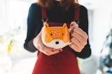 A person holds a large ceramic mug designed to resemble the face of a fox, with orange and white colors contrasting against the person's dark clothing. Soft lighting creates a warm atmosphere, while the focus is on the mug, highlighting its whimsical design.