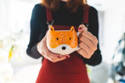 A person holds a large ceramic mug designed to resemble the face of a fox, with orange and white colors contrasting against the person's dark clothing. Soft lighting creates a warm atmosphere, while the focus is on the mug, highlighting its whimsical design.