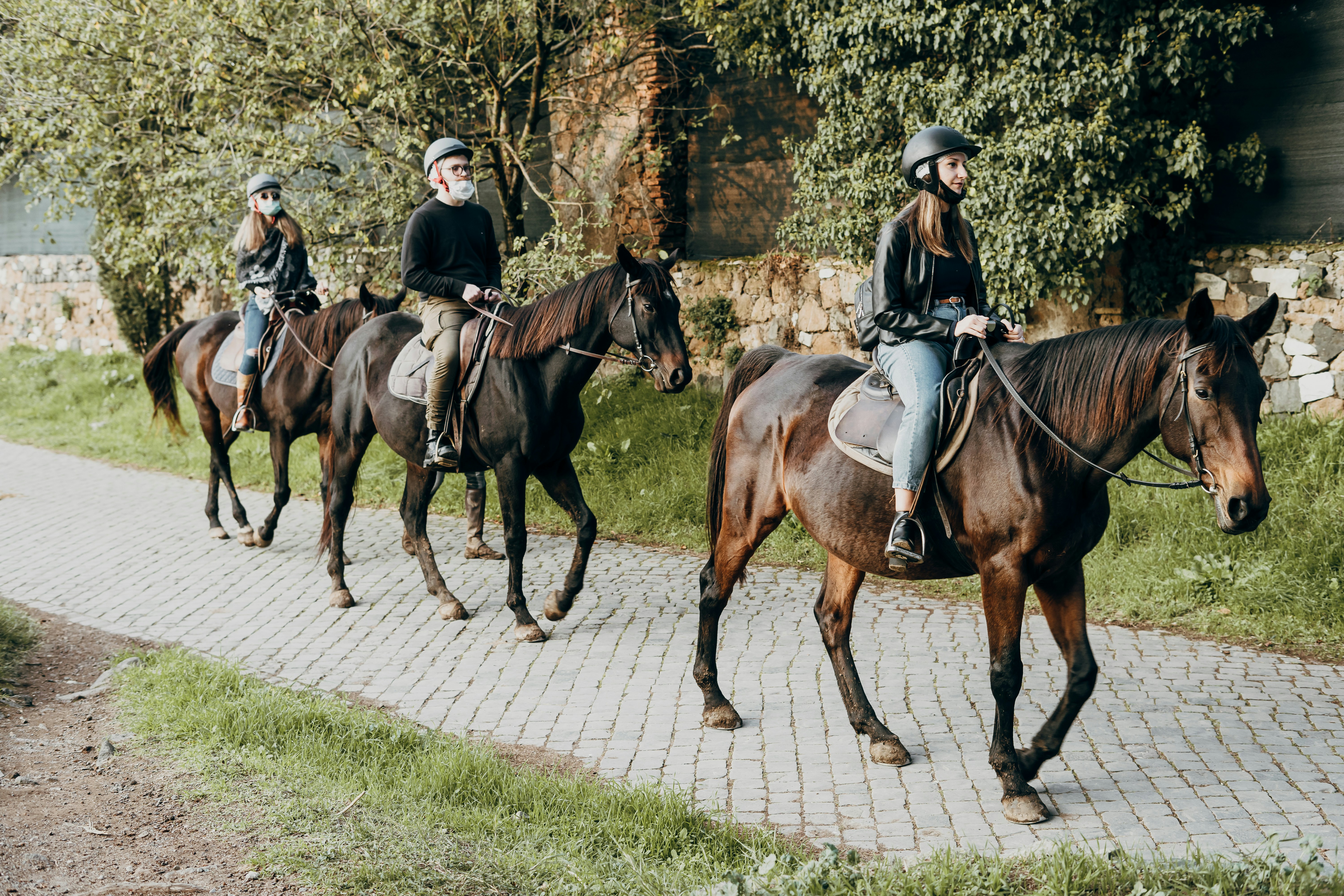man riding horse on green grass field during daytime, Two women and a man, wearing face masks, ride horses along via Appia Antica in Rome
