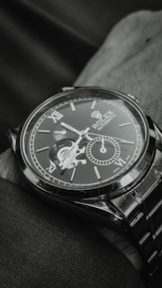A close-up view of an elegant wristwatch with a visible mechanical movement. The dial includes Roman numerals and a smaller sub-dial, creating a sophisticated and detailed look. The watch is resting on a fabric surface, and the image is captured in a monochromatic tone.