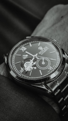 A close-up view of an elegant wristwatch with a visible mechanical movement. The dial includes Roman numerals and a smaller sub-dial, creating a sophisticated and detailed look. The watch is resting on a fabric surface, and the image is captured in a monochromatic tone.