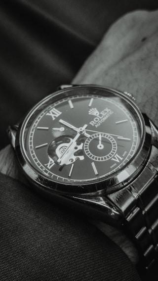 A close-up view of an elegant wristwatch with a visible mechanical movement. The dial includes Roman numerals and a smaller sub-dial, creating a sophisticated and detailed look. The watch is resting on a fabric surface, and the image is captured in a monochromatic tone.