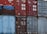 A stack of old shipping containers in various colors and conditions, with visible logos, numbers, and text on their surfaces. The containers are primarily red and blue, each showing signs of weathering and rust.