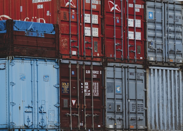 A stack of old shipping containers in various colors and conditions, with visible logos, numbers, and text on their surfaces. The containers are primarily red and blue, each showing signs of weathering and rust.