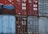 A stack of old shipping containers in various colors and conditions, with visible logos, numbers, and text on their surfaces. The containers are primarily red and blue, each showing signs of weathering and rust.