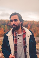 A man is standing outdoors, dressed in a layered outfit that includes a plaid shirt and a jacket with a furry lining. He is in a natural setting with a background of autumnal trees showing vibrant fall colors.