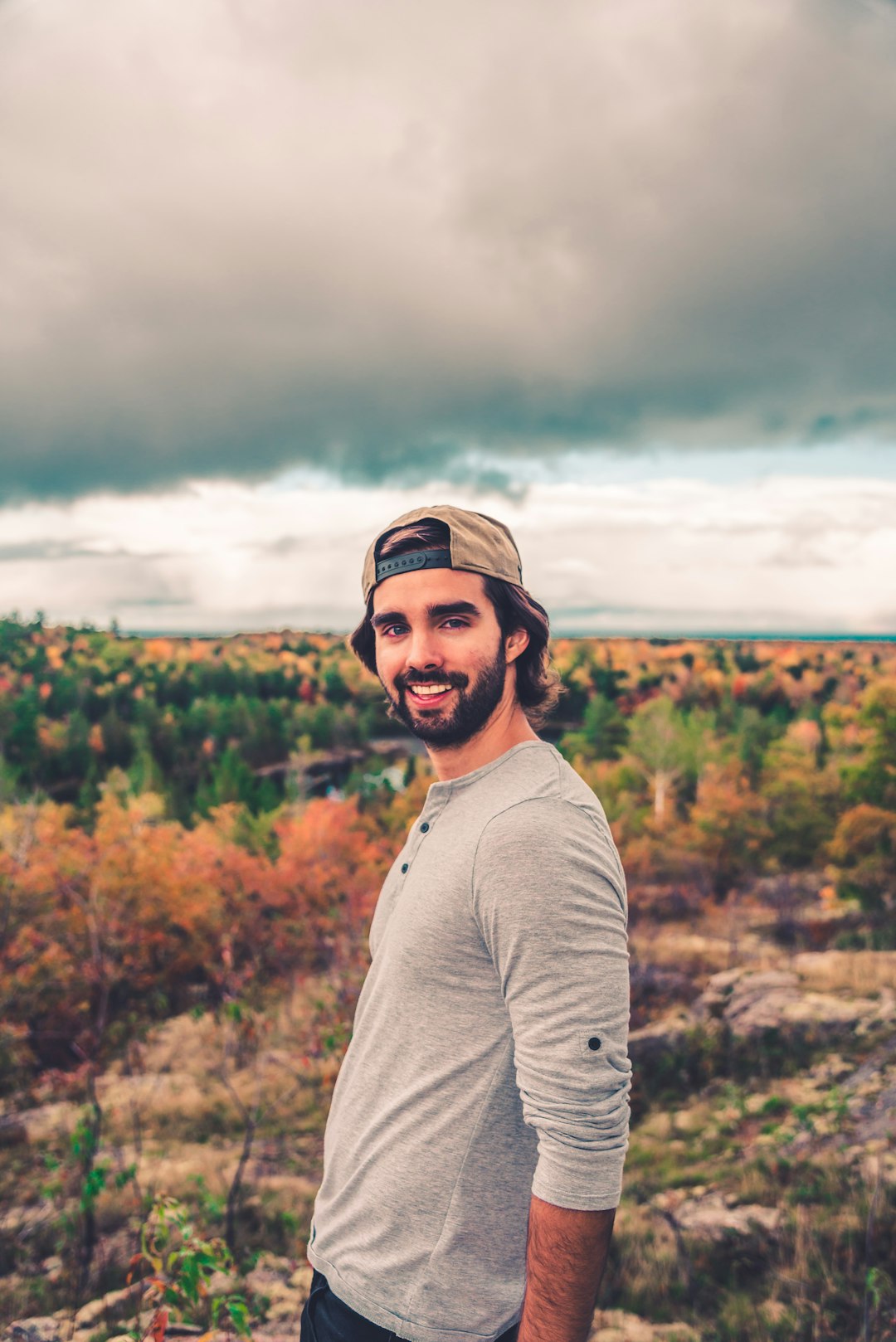 man in gray long sleeve shirt standing near green trees under white clouds during daytime