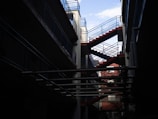 A modern architectural design featuring an exterior staircase with red steps and blue railings. The structure is set between buildings, with a partially visible sky and pipes running horizontally across the foreground.