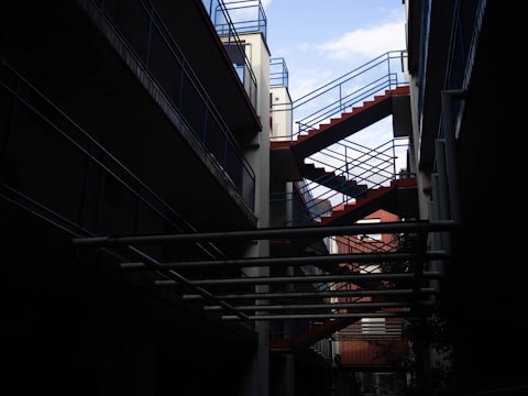 A modern architectural design featuring an exterior staircase with red steps and blue railings. The structure is set between buildings, with a partially visible sky and pipes running horizontally across the foreground.