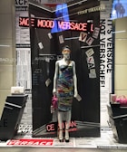 A mannequin dressed in a vibrant, multicolored dress, displayed in a high-fashion store window. The background features newspaper-like prints and various texts. Around the mannequin are luxury handbags, one white and quilted, the other pink, adding to the upscale theme. An LED sign displays the word 'VERSACE' in red lights.