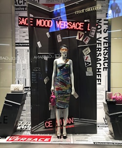 A mannequin dressed in a vibrant, multicolored dress, displayed in a high-fashion store window. The background features newspaper-like prints and various texts. Around the mannequin are luxury handbags, one white and quilted, the other pink, adding to the upscale theme. An LED sign displays the word 'VERSACE' in red lights.