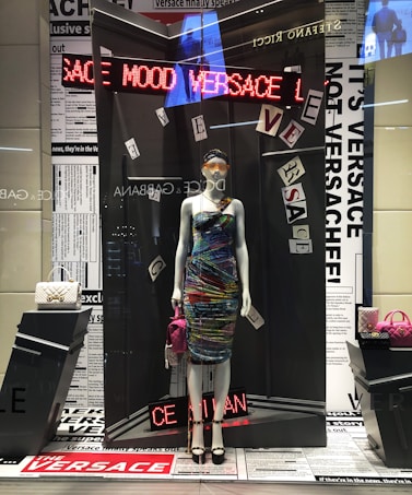 A mannequin dressed in a vibrant, multicolored dress, displayed in a high-fashion store window. The background features newspaper-like prints and various texts. Around the mannequin are luxury handbags, one white and quilted, the other pink, adding to the upscale theme. An LED sign displays the word 'VERSACE' in red lights.