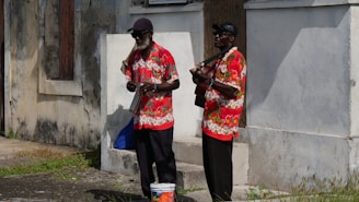 Two older men wearing colorful floral shirts, black hats, and dark trousers are playing musical instruments on a sunny street corner. The backdrop consists of an aged stucco wall, adding a rustic vibe to the scene. One man is holding a small banjo, while the other plays a guitar.
