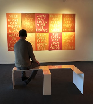 A person sitting on a modern, minimalist bench in a gallery setting, facing a series of nine square canvases on the wall. Each canvas displays text with provocative messages in bold capital letters. The canvases feature a gradient of warm colors ranging from deep red to orange and yellow.
