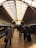 A bustling train station with people walking along the tiled floor towards the trains. The wide arched ceiling has a pattern of beams and glass panels, allowing natural light to illuminate the station. To the left, a train is stationed, featuring vibrant colors, while travelers carry bags and suitcases, creating a sense of movement and travel.