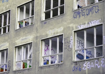 A building facade with multiple broken windows, each surrounded by vibrant graffiti art. The walls have a weathered appearance, accentuated by faded and peeling paint. The interior of the rooms visible through the windows also displays various graffiti tags and designs.