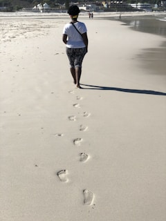 A person walking slowly along a sandy beach, footprints trailing behind.