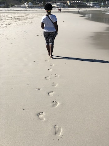 A person walking slowly along a sandy beach, footprints trailing behind.