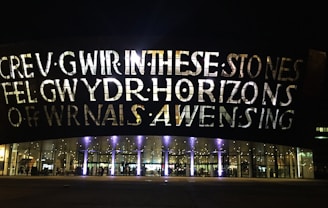 A large building facade is illuminated by lights, showcasing large bold letters that spell out text. The building has a series of arched windows at the lower level, through which interior lighting and reflections are visible. The overall scene is set during nighttime, adding to the dramatic effect of the illuminated text and glass.