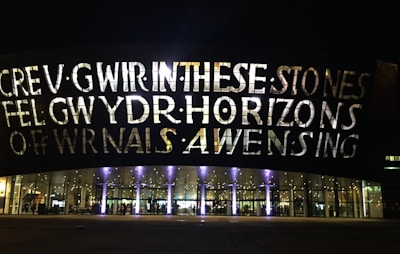 A large building facade is illuminated by lights, showcasing large bold letters that spell out text. The building has a series of arched windows at the lower level, through which interior lighting and reflections are visible. The overall scene is set during nighttime, adding to the dramatic effect of the illuminated text and glass.