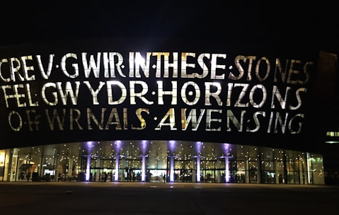 A large building facade is illuminated by lights, showcasing large bold letters that spell out text. The building has a series of arched windows at the lower level, through which interior lighting and reflections are visible. The overall scene is set during nighttime, adding to the dramatic effect of the illuminated text and glass.